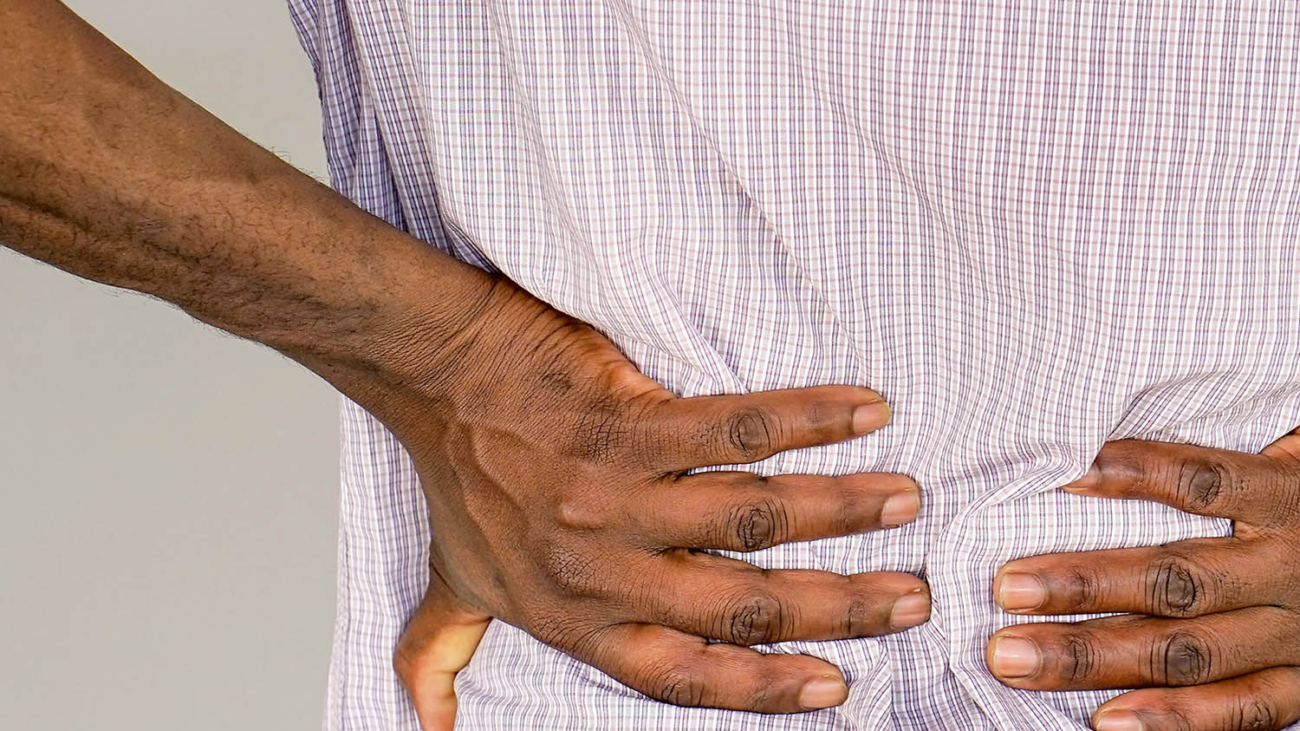 Closeup of african man with pain in kidneys on gray background. Young man with back ache clasping her hand to her lower back. Man suffering from ribbing pain, waist pain.